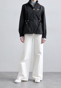 Person som bär en svart zip-up jacka med EAZ-logotyp, vita vida byxor, ljusgrå handskar och vita sneakers står på grått golv.