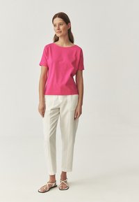 Top de manga corta, de color rosa brillante, con un escote amplio, combinado con pantalones blancos ajustados. La modelo lleva sandalias planas blancas con tiras.