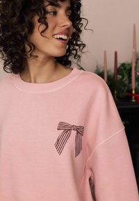 Jasnoróżowy sweatshirt z okrągłym dekoltem, z czarnym paskowanym wzorem w kształcie kokardy po lewej stronie klatki piersiowej. Miękka tkanina i luźny krój.