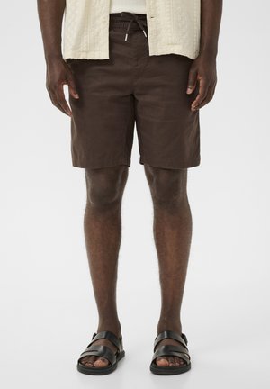 Egy férfi sötétbarna, térdig érő rövidnadrágot, fekete bőrszandált és világos krémszínű, texturált rövid ujjú inget visel, előre nézve áll.