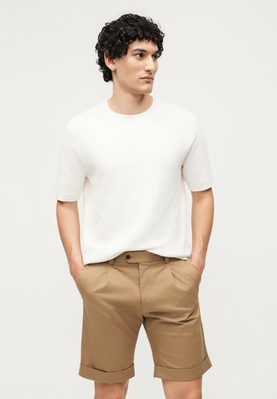 Άνδρας με καστανά σγουρά μαλλιά που φοράει άσπρο κοντομάνικο πουκάμισο και καφέ σορτς που είναι γυρισμένα, με τα χέρια στις τσέπες, κοιτάζοντας πλάγια σε απλό φόντο.