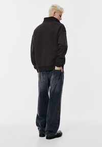 Svart sweatshirt med hög krage, avslappnad passform, ribbade muddar, kombinerad med mörka, randiga, vida jeans och svart fotbeklädnad. Minimalistisk design, enfärgad bakgrund.