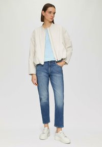 Veste bomber beige clair, t-shirt bleu clair, jeans bleu moyen et baskets à plateforme blanches. Matière lisse, coupe décontractée, jeans longueur cheville.