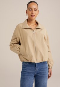 Beige bomber bunda se zipovým zapínáním, širokým límcem a elastickými manžetami; kombinováno s modrými džíny s předními kapsami. Hladká textilie.