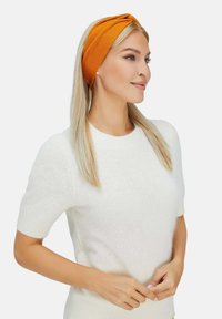 Fascia in maglia arancione con design frontale attorcigliato, indossata da una persona in un maglione bianco soffice a maniche corte, con lunghi capelli biondi.