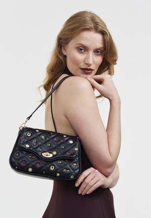 Žena svijetlosmeđe kose u tamnoj haljini bez rukava drži crnu prošivenu torbicu ukrašenu šarenim izvezenim simbolima.