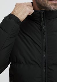 Črna podložena jakna z visokim ovratnikom in zadrgo. Ima elastične manšete in sijočo teksturo blaga.