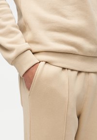 Μπεζ φούτερ με ριμπ μανσέτες, σετ με αντίστοιχα τζόγκερ με πλαϊνές τσέπες και χαλαρή εφαρμογή, κατασκευασμένα από απαλό, υφασμάτινο ύφασμα με υφή.