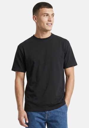 Ung mand med kort hår iført en simpel sort t-shirt med korte ærmer og blå jeans, kigger mod højre, hænderne i lommerne.