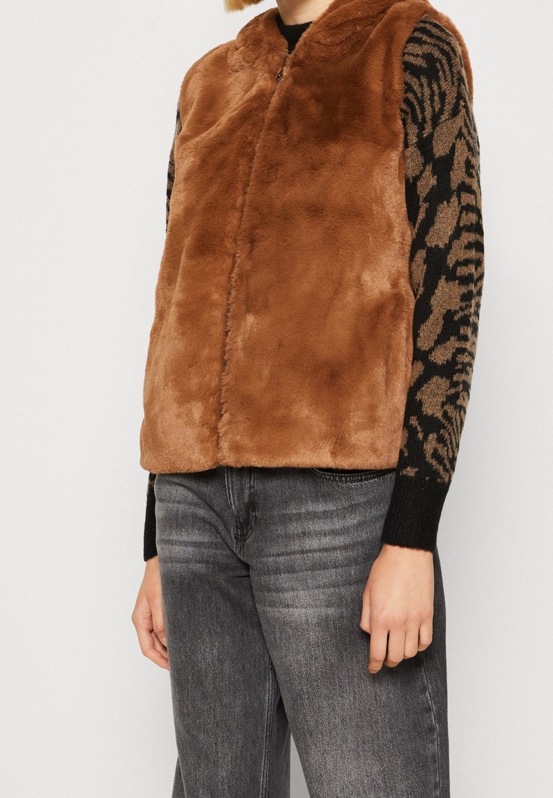 Personne portant une veste en fausse fourrure marron par-dessus un pull à manches longues imprimé animal noir et marron, associée à un jean noir délavé.