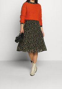 Tenue superposée comprenant un pull en maille orange vif et une robe à imprimé floral noir. Les accessoires incluent un sac à main noir et des bottines crème.