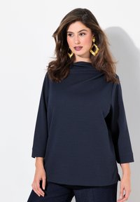 Top blu navy con scollatura drappeggiata, maniche ¾, tessuto texturizzato e vestibilità ampia, abbinato a grandi orecchini geometrici in oro.