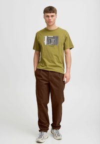 Olivgrünes Baumwoll-T-Shirt mit einem schwarz-weißen Grafikdruck, kombiniert mit braunen Hosen. Die Schuhe sind grau und beige Sportschuhe.
