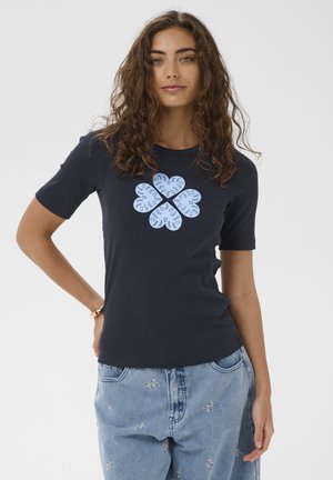 Giovane donna con capelli ricci castani che indossa una maglietta blu navy con simboli a forma di cuore blu e jeans blu chiaro ricamati, in piedi con la mano sul fianco.