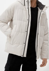 Homme portant une doudoune beige clair par-dessus une chemise noire et un pantalon cargo noir, une main dans la poche.