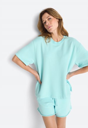 Vrouw met lichtbruin haar, gekleed in een loszittend lichtblauw kort mouwtopje en bijpassende korte broek, staand met handen in de zij tegen een witte achtergrond.