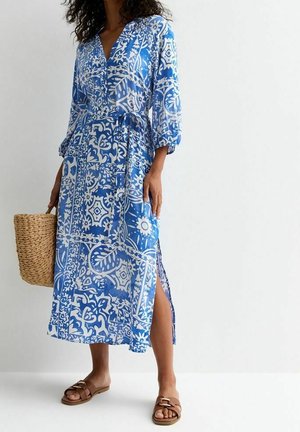 Kobieta w niebiesko-białej midi sukience w wzory z rozcięciem z boku, trzyma pleciony kosz i nosi brązowe sandały na płaskiej podeszwie.