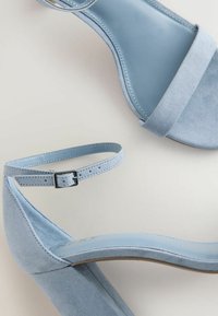 Sandales à talons hauts en daim bleu clair avec brides à la cheville et boucles argentées sur un fond uni clair.