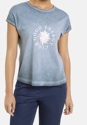 Persoon die een blauw T-shirt met korte mouwen draagt met "GOOD ENERGY CLUB" en zonafbeelding, gecombineerd met donkerblauwe broek, torso en armen zichtbaar.