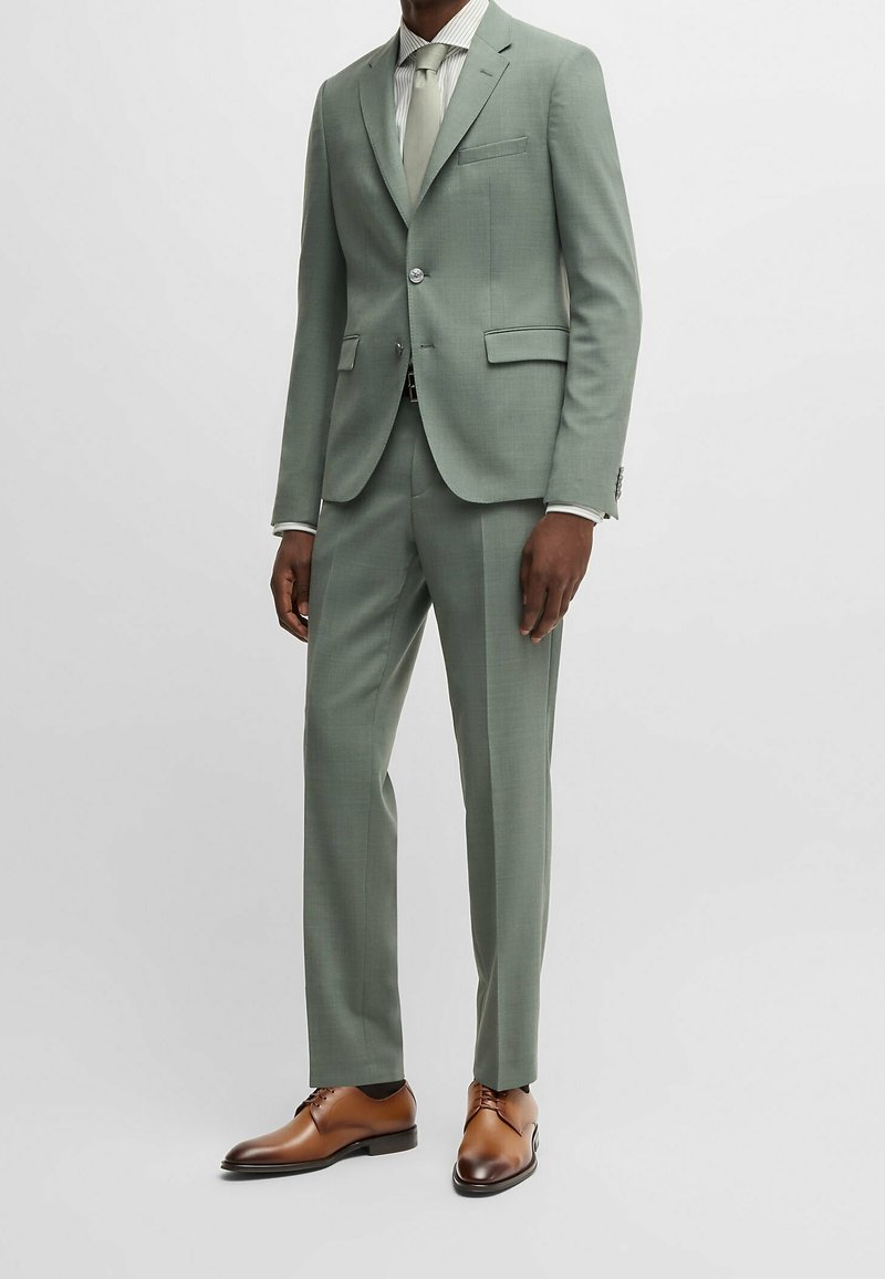 Homme portant un costume vert clair avec un pantalon assorti, une chemise blanche, une cravate vert pâle et des chaussures habillées en cuir marron sur un fond uni.