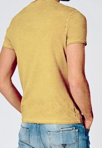 Mand iført en sennepsgul T-shirt og lyseblå denimjeans, står med hænderne i lommerne og vender bort.