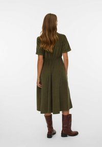 Vestido verde oliva con mangas cortas, cintura ajustada y falda fluida. Combinado con botas marrones hasta la rodilla. Textura suave, diseño casual.
