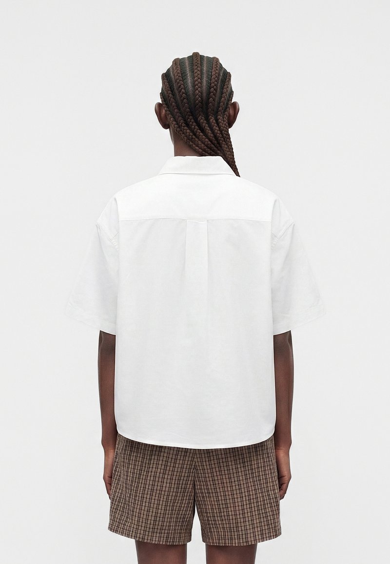 Person with long braided hair wearing a loose white short-sleeve shirt and plaid shorts, shown from the back against a plain background.