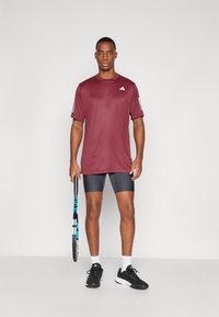 Camisa atlética marrón de manga corta, combinada con pantalones cortos gris oscuro. Zapatillas deportivas negras y calcetas blancas. Sosteniendo una raqueta de tenis.