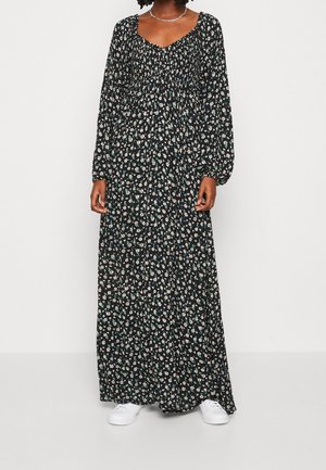 Frau trägt ein schwarzes Maxikleid mit langen Ärmeln mit kleinen weißen und grünen Blumenmustern und weißen Turnschuhen vor einem einfarbigen Hintergrund.