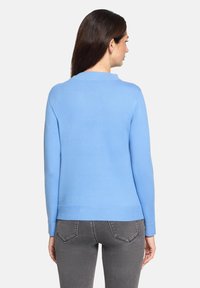 Kobieta o długich brązowych włosach, ubrana w jasnoniebieski sweter i ciemnoszare dżinsy, stojąca tyłem na jednolitym tle.