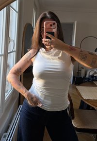 Top bianco senza maniche con scollo drappeggiato, indossato con jeans scuri. Tatuaggi evidenti sulle braccia, con una texture morbida e liscia.