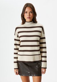 Sweter w paski w kolorze kremowym i brązowym, z wysokim kołnierzem, prążkowanymi mankietami i luźnym krojem. Połączony z czarną mini spódnicą.