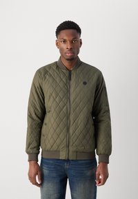 Veste en bomber matelassée de couleur vert olive. Dotée d'une fermeture éclair à l'avant, de poignets côtelés et de deux poches latérales. Texture lisse avec un léger éclat.