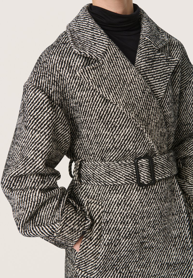 Abrigo de mezcla de lana en patrón de espiga en blanco y negro, con cuello de chal, mangas anchas, cinturón ajustable y bolsillos laterales.