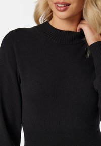 Pull en tricot noir à côtes, présentant un col montant et une coupe ajustée. La texture semble douce et lisse, sans motifs ni accents visibles.
