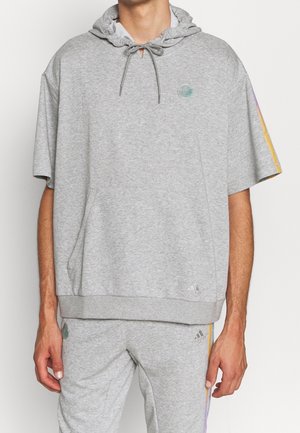 Mann trägt hellgrauen Kapuzenpullover mit kurzen Ärmeln und passende Jogginghose mit kleinen Adidas-Logos und mehrfarbigen Streifen an Ärmeln und Hosenbeinen.