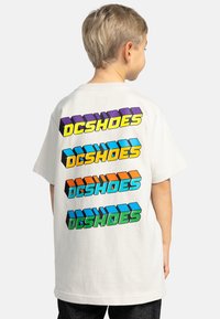 Bílý bavlněný tričko s barevným grafickým textem "DCSHOES" v různých barvách a 3D efekty na zadní straně. Standardní střih, krátké rukávy.