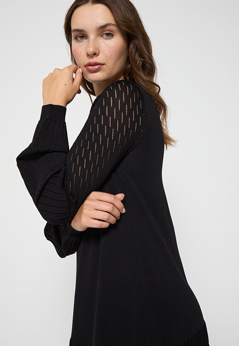 Pull noir avec texture côtelée et manches transparentes à motif. Design ajusté avec un col rond et un léger volant à l'ourlet.