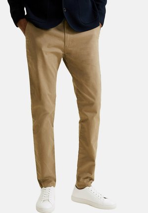Homme portant un chino beige slim-fit avec les mains dans les poches, associé à des baskets blanches et un blazer foncé, debout sur un fond uni.