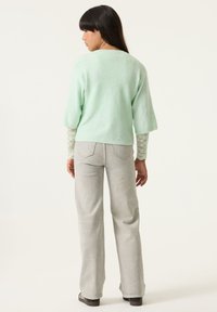 Suéter de manga corta color verde menta con acabado texturizado, superpuesto sobre una camisa de encaje de manga larga. Jeans acampanados grises y zapatos oscuros completan el conjunto.