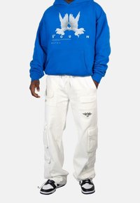 Modrá mikina s kapucí a grafikou bílé holubice s textem, spárovaná s bílými cargo kalhotami s bočními kapsami a černým logem, a černými teniskami.