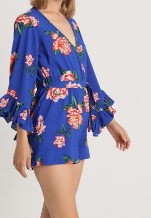 Kobieta ubrana w niebieską, kwiecistą sukienkę kopertową z falbanami na rękawach 3/4 i paskiem w talii, stojąca na jednolitym jasnym tle.