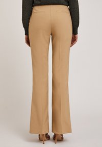 Pantalons évasés beiges avec une texture lisse, dotés de deux poches arrière et d'une taille mi-haute, associés à des sandales à talons hauts.