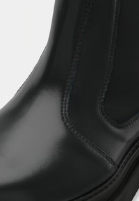 Bota de tornozelo em pele preta com acabamento brilhante, apresentando costuras curvas e um painel lateral elástico texturizado. Acentos em costura azul.