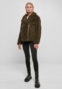 Bruine, fluffy fleece jas met een brede kraag en gouden rits, gecombineerd met zwarte strakke jeans en stevige zwarte laarzen.