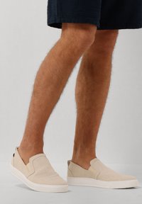 Beige instappers met een textiel bovenwerk en elastische zijkanten, rustend op een witte rubberen zool. Minimalistisch ontwerp met afgeronde randen.