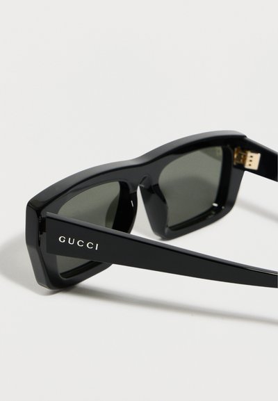Μαύρα ορθογώνια γυαλιά ηλίου με σκούρους φακούς, που φέρουν το λογότυπο της Gucci στο βραχίονα του πλαισίου, σε λευκό φόντο.