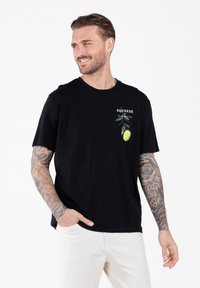 Man met getatoeëerde armen, gekleed in een zwarte t-shirt met een citroenafbeelding en de tekst "Squeeze La Giornata", gecombineerd met witte broek.