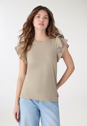 Giovane donna con capelli castani ondulati che indossa un top beige senza maniche con maniche arricciate a occhiello e jeans azzurri, in piedi davanti a uno sfondo bianco.