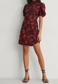 Modellen poserar i en bordeaux blommig klänning med puffärmar, kombinerad med svarta remsandaler, vilket visar en avslappnad men stilfull look.